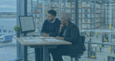 HR director and recruitment manager reviewing candidates at a logistics office overlooking a warehouse, illustrating AI recruitment.