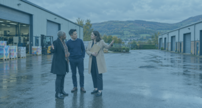 Two company co-owners and their adviser discussing a demerger or shareholder buyout in a Cardiff print factory yard, with separate industrial units opposite.